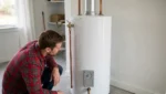 What That Water Heater Noise Means Homeowner Kneeling Beside A Tall Tank Water Heater In A Utility Room, Listening To The Tank.